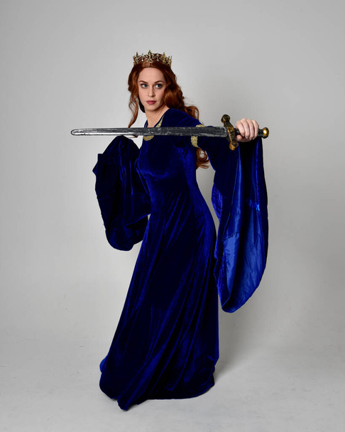 Full length portrait of  girl wearing long blue velvet gown with golden crown, holding a sword. standing pose  isolated against a studio background. - Fotoğraf, Görsel
