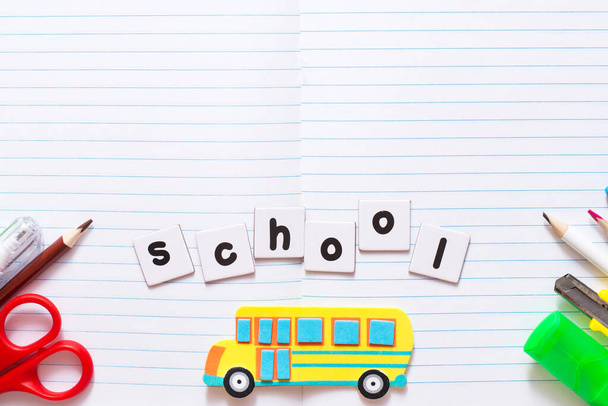 Текст ШКОЛА, написанный печатными буквами на белых листах бумаги на листе блокнота с красочной школьной канцелярской принадлежностью и желтым игрушечным школьным автобусом - Фото, изображение