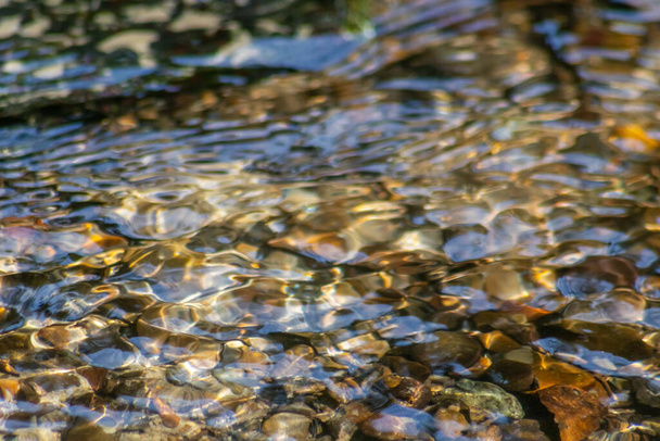 クリスタルクリアウォータークリークの水の中に日当たりの良い反射を持つ輝く水の中の石は、牧歌的な自然背景として禅の瞑想、小さな波と健康な山の春に絹のような波紋を示しています - 写真・画像
