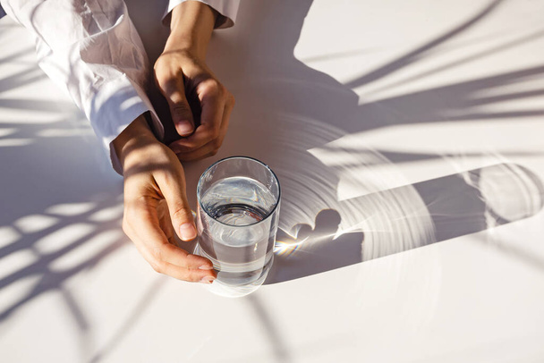 Woman's hands and a glass of clean water on the white table in natural sunlight with plant shadows. Minimalist lifestyle concept - Photo, image