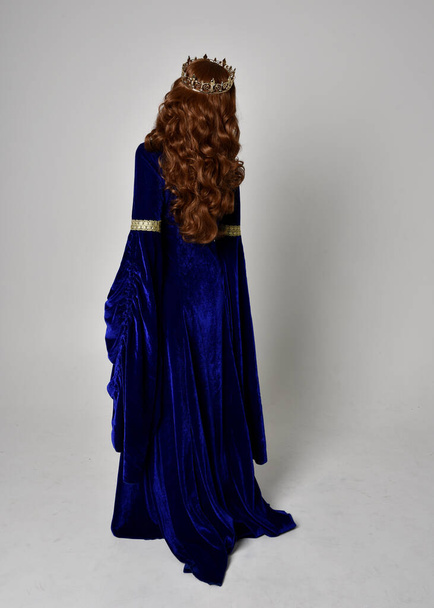 Full length portrait of  girl wearing long blue velvet gown with golden crown. standing pose with back to the camera, isolated against a studio background. - Photo, Image
