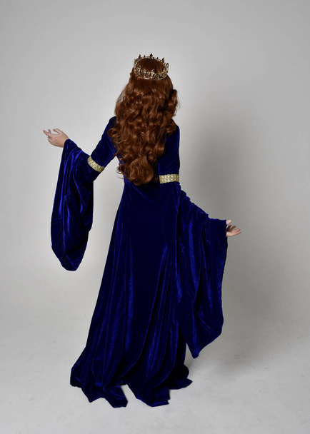 Full length portrait of  girl wearing long blue velvet gown with golden crown. standing pose with back to the camera, isolated against a studio background. - 写真・画像