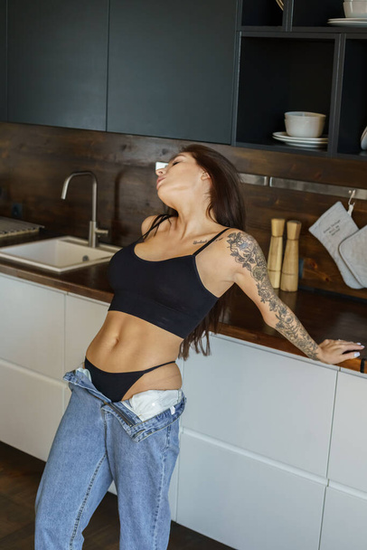 Sporty brunette woman with tattoos on hand and on body posing on kitchen. Portrait of young woman wearing blue jeans and black top standing at apartments interior  - Foto, Imagen