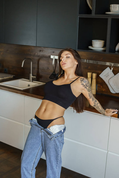 Sporty brunette woman with tattoos on hand and on body posing on kitchen. Portrait of young woman wearing blue jeans and black top standing at apartments interior  - Foto, afbeelding