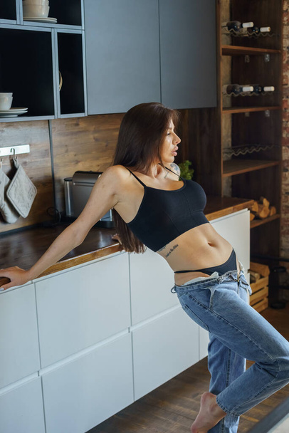 Sporty brunette woman with tattoos on hand and on body posing on kitchen. Portrait of young woman wearing blue jeans and black top standing at apartments interior  - Photo, image