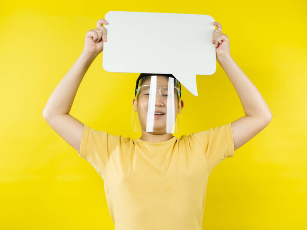 Asian preteen boy wearing face shield holding blank speech bubble on yellow background. - Zdjęcie, obraz
