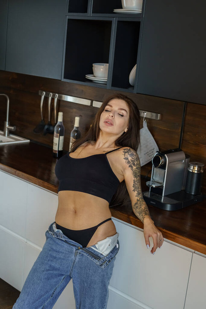Sporty brunette woman with tattoo on hand posing on kitchen. Portrait of young woman wearing blue jeans and black top standing at apartments interior  - Photo, Image