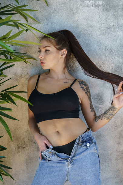 Brunette woman with tattoos on body posing on gray wall background. Portrait of young woman wearing blue jeans and black top standing at apartments interior  - Valokuva, kuva