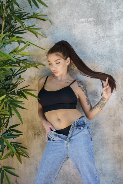 Brunette woman with tattoos on body posing on gray wall background. Portrait of young woman wearing blue jeans and black top standing at apartments interior  - Foto, immagini