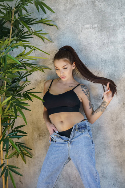 Brunette woman with tattoos on body posing on gray wall background. Portrait of young woman wearing blue jeans and black top standing at apartments interior  - Foto, Bild