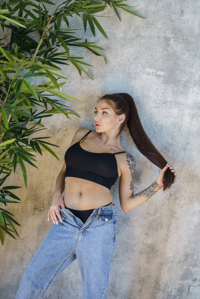 Brunette woman with tattoos on body posing on gray wall background. Portrait of young woman wearing blue jeans and black top standing at apartments interior  - Foto, Imagen