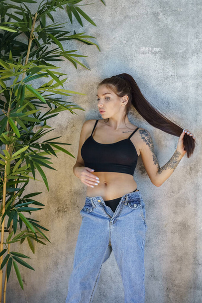 Brunette woman with tattoos on body posing on gray wall background. Portrait of young woman wearing blue jeans and black top standing at apartments interior  - Photo, image