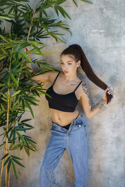 Brunette woman with tattoos on body posing on gray wall background. Portrait of young woman wearing blue jeans and black top standing at apartments interior  - 写真・画像