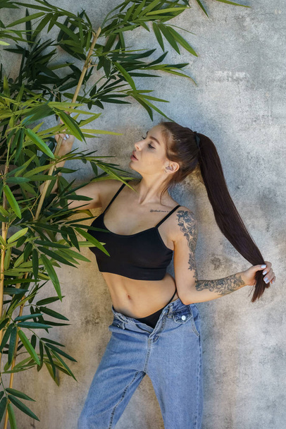 Brunette woman with tattoos on body posing on gray wall background. Portrait of young woman wearing blue jeans and black top standing at apartments interior  - Foto, imagen