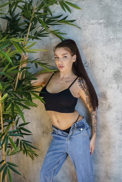 Brunette woman with tattoos on body posing on gray wall background. Portrait of young woman wearing blue jeans and black top standing at apartments interior  - Photo, Image