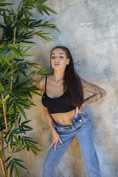 Brunette woman with tattoos on body posing on gray wall background. Portrait of young woman wearing blue jeans and black top standing at apartments interior  - Φωτογραφία, εικόνα