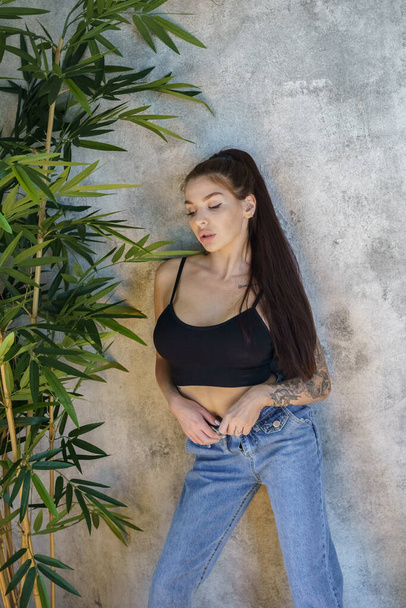 Brunette woman with tattoos on body posing on gray wall background. Portrait of young woman wearing blue jeans and black top standing at apartments interior  - Foto, imagen