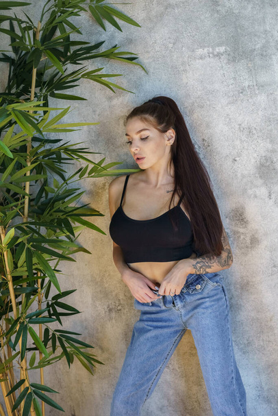 Brunette woman with tattoos on body posing on gray wall background. Portrait of young woman wearing blue jeans and black top standing at apartments interior  - Photo, image