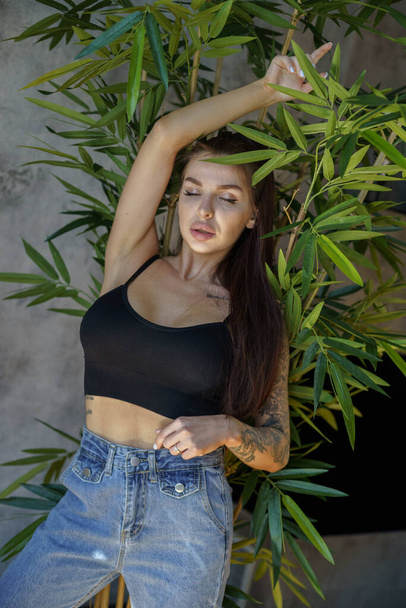Brunette woman with tattoos on body posing on gray wall background. Portrait of young woman wearing blue jeans and black top standing at apartments interior  - Foto, immagini