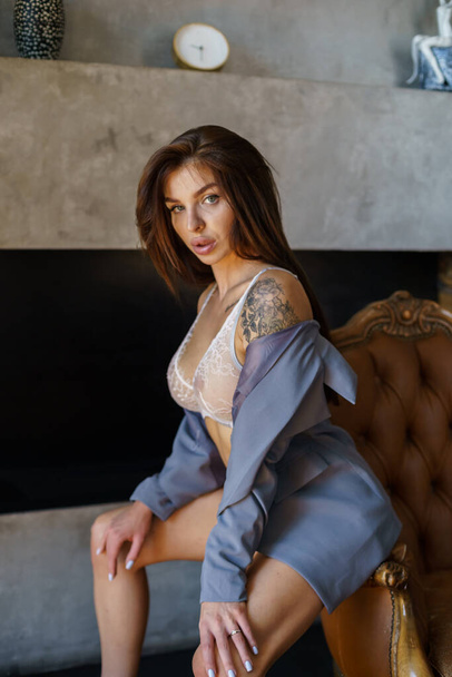 Sexual brunette model with tattoos on body posing at luxury apartments interior. Studio portrait of young woman wearing white lingerie and blue jacket  - 写真・画像