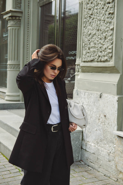 Outdoor fashion portrait of a  young woman. Streetstyle. A brunette on the streets of the city in a fashionable oversized jacket and sunglasses from the sun. - 写真・画像