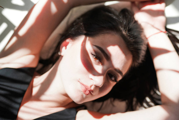 Portrait de beauté du visage féminin à la peau naturelle, femme allongée sur fond blanc ensoleillé en sous-vêtements noirs - Photo, image