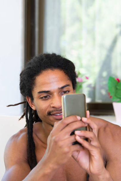 Gömleksiz yakışıklı genç siyah adam gülümsüyor ve arka planda telefonuna bakıyor. İletişim ve eğlence kavramı. - Fotoğraf, Görsel