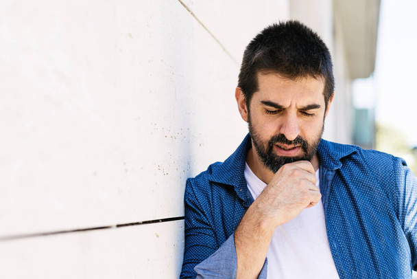 Задумчивый бородатый мужчина опирается на стену с рукой на подбородке - Фото, изображение