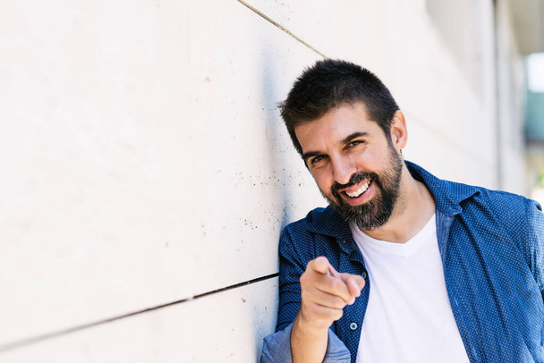 Bearded smiling male leaning on wall while pointing to you with finger - Photo, Image
