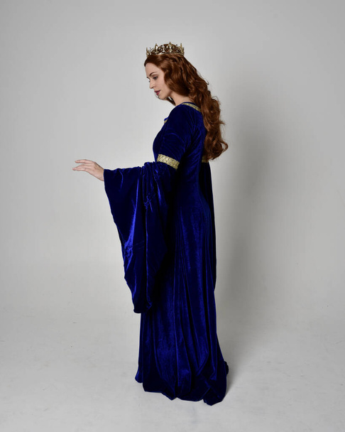 Full length portrait of  girl wearing long blue velvet gown with golden crown. standing pose with back to the camera, isolated against a studio background. - Photo, Image