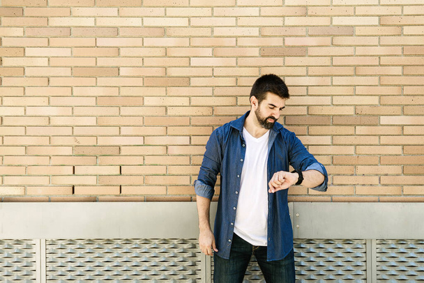 baard man kijken op zijn horloge tegen een baksteen muur buiten - Foto, afbeelding