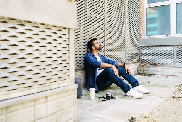 Homme barbu joyeux en tenue décontractée assis sur le sol de la rue tout en se relaxant sur une pause café - Photo, image