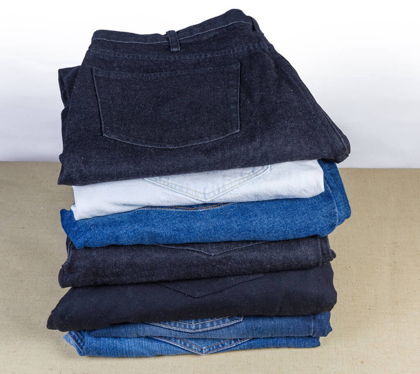 Несколько различных пар джинсов различных цветов сложены в стеке на текстильной поверхности - Фото, изображение