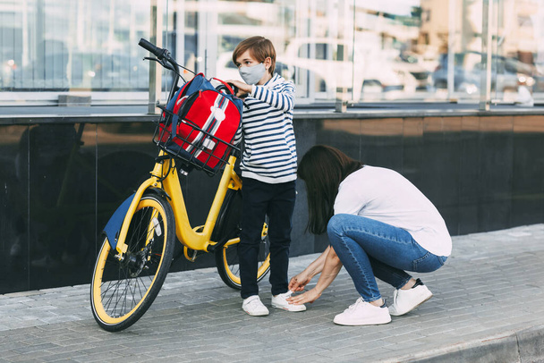 Egy anya beállítja a fia tornacipőjét, mielőtt biciklizne az iskolába. Egy fiú maszkot visel, és hátizsákot cipel, hamarosan iskolába megy a koronavírus járvány idején.. - Fotó, kép