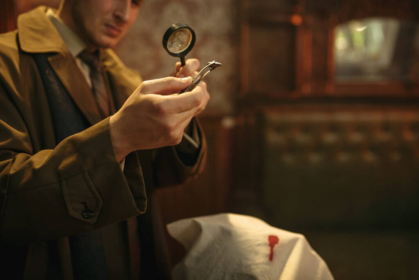 Male detective looks on evidence through magnifying glass at the crime scene, retro style. Criminal investigation, inspector is working on a murder, vintage room interior on background - Photo, Image