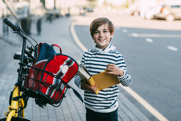 Veselý chlapec drží knihu v rukou a stojí vedle kola s batohem zavěšeným na řídítka. - Fotografie, Obrázek