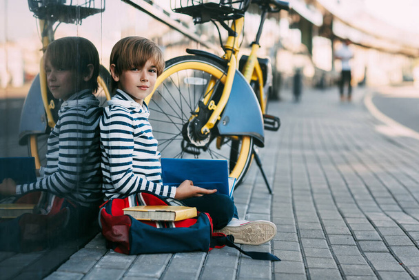 Un lindo colegial está sentado al aire libre en la escuela y sosteniendo un libro en sus manos. Junto a ella hay una mochila escolar y una bicicleta
 - Foto, imagen