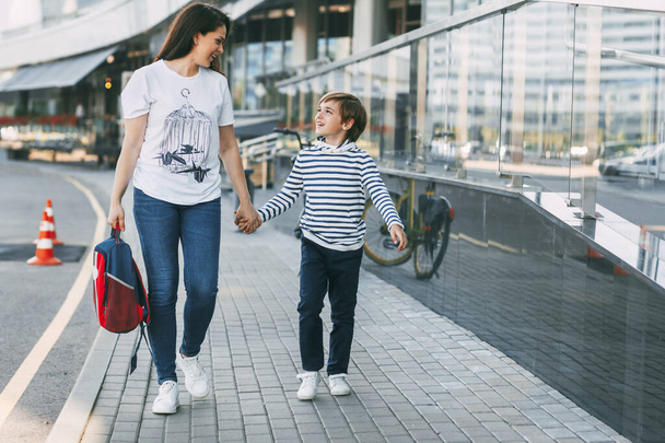 The mother carries a backpack in one hand, and holds her son by the other hand. A cheerful smiling boy walks with his mother from school, space for text - Foto, imagen