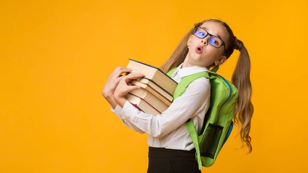 Niña de la escuela con exceso de trabajo sosteniendo libros de pie sobre fondo amarillo, Panorama
 - Foto, imagen