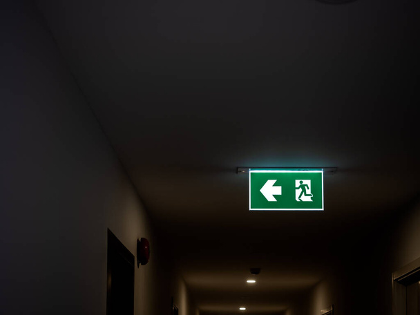 A vészkijárat. Világító zöld vészkijárat tábla lóg a mennyezeten a modern épületben a sötétben lakásban fénymásoló hely. - Fotó, kép