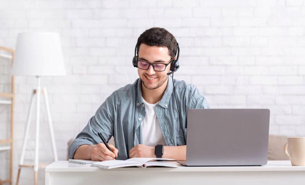 遠隔教育と独立教育。ヘッドフォンを持つ笑顔の男は、デスクトップ上のノートパソコンとノートブックでノートを作ります - 写真・画像