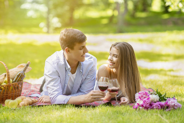 Romanic girlfriend with her boyfriend lying on picnic blanket and drinking wine at park on summer day - Photo, Image