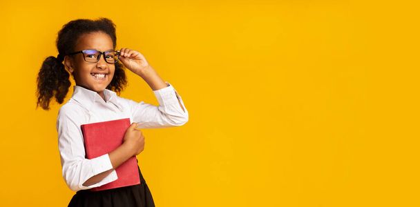 Pequeña colegiala africana sosteniendo libro sonriendo ajustando gafas, fondo amarillo
 - Foto, Imagen