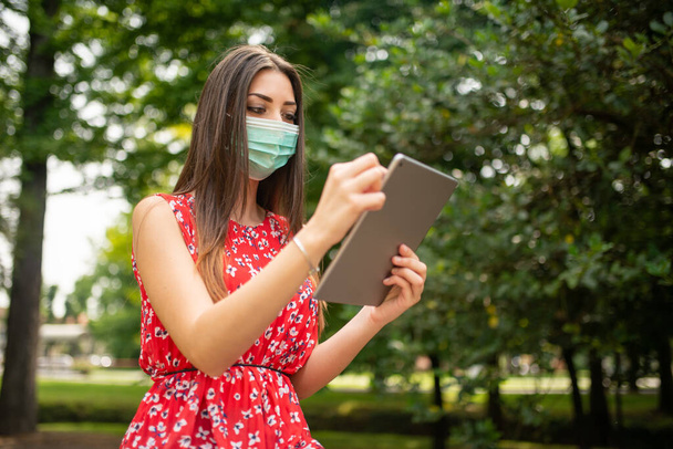 Masked young woman using a tablet in the park - Foto, Bild