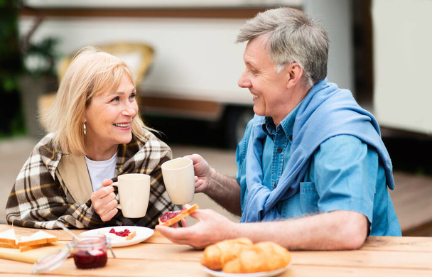 Cheerful mature man and his wife enjoying morning coffee with jam toasts at campground - Foto, imagen