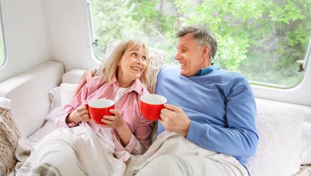 Senior married couple with cups of hot coffee lying in bed inside of their camping vehicle - Photo, Image