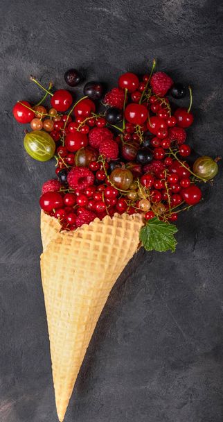 An explosion of different berries. Photo of blueberries, strawberries, raspberries in a waffle cone on a black textured background. View from above. - Photo, Image