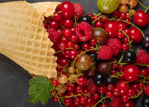 An explosion of different berries. Photo of blueberries, strawberries, raspberries in a waffle cone on a black textured background. View from above. - Фото, зображення