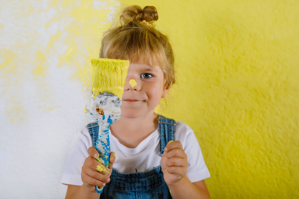 Χαριτωμένο κοριτσάκι ζωγραφική στον τοίχο με κίτρινο χρώμα στο νέο σπίτι. Οικογενειακό σπίτι επισκευών. Ευτυχισμένο κορίτσι ζωγραφίζει τον τοίχο, επιλέγοντας χρώμα με παλέτα, διασκεδάζοντας με πινέλο, σε εσωτερικούς χώρους - Φωτογραφία, εικόνα