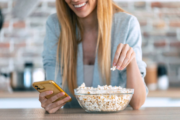 Shot of smiling woman grabbing and picking popcorn from a bowl to eat while using her mobile phone at home. - Valokuva, kuva